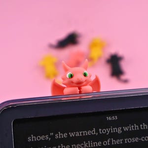 Op de afbeelding: Een roze drakenfiguur met groene ogen en rode vleugels zit op een zwart elektronisch apparaat. De draak is het middelpunt, met andere kleurrijke vormen wazig op de achtergrond tegen een roze achtergrond. De tijd 16:53 is zichtbaar.