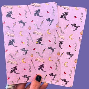 May include: Two pink sheets of paper with a repeating pattern of black dragons, swords, and open books. The background is a light pink with a subtle glitter effect.