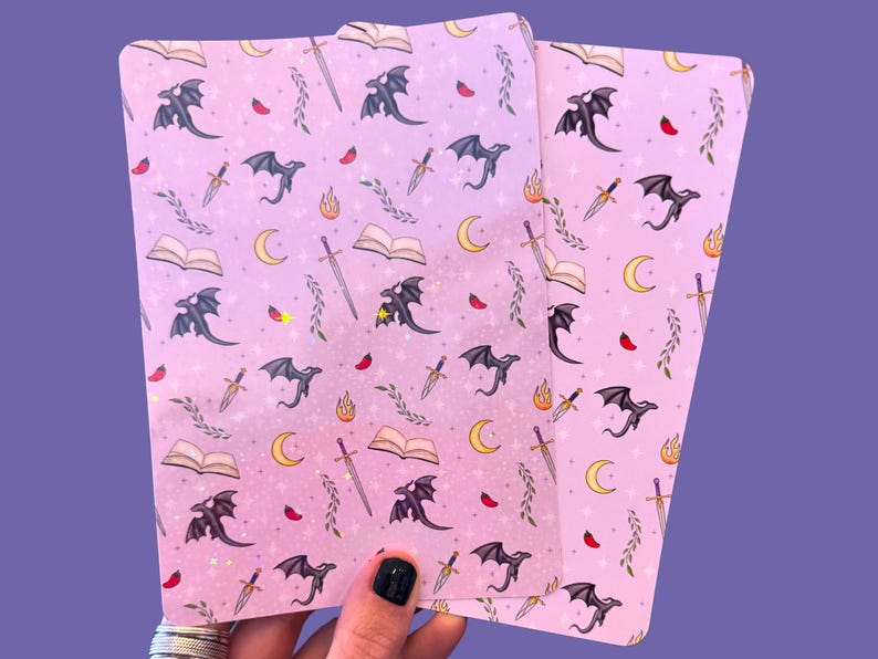 May include: Two pink sheets of paper with a repeating pattern of black dragons, swords, and open books. The background is a light pink with a subtle glitter effect.