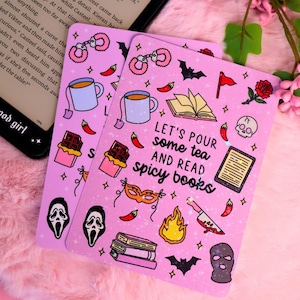 May include: Pink rectangular card featuring illustrations of tea cups, chilli peppers, books, and a skull. The card reads "LET'S POUR some tea AND READ spicy books". A black e-reader is visible in the background.