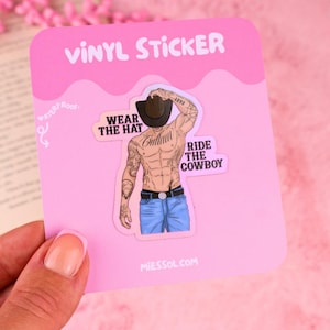 Op de afbeelding: Een vinylsticker op een roze achtergrondkaart met een getatoeëerd persoon zonder shirt, met een bruine cowboyhoed en een blauwe spijkerbroek. De tekst luidt "WEAR THE HAT" en "RIDE THE COWBOY" met "1990" op de hoed. De sticker is gemarkeerd als "WATERPROOF".