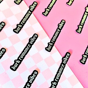 May include: A collection of stickers with the text "dark romance vibes" in a gothic font. The stickers are on a pink and white checkered background.