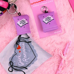 Op de afbeelding: Een paarse schermreinigingsdoek-sleutelhanger met de tekst "reading something unholy" op een boekafbeelding. De doek is bevestigd aan een zilveren sleutelhanger. Een roze pen en een "Dark Romance" doek zijn ook zichtbaar.