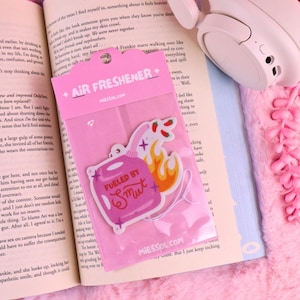 May include: A pink air freshener featuring a cartoon gas can and flames, with the words "Fueled by Smut." The item is packaged in clear plastic, labelled "AIR FRESHENER" and "MIESSOL.COM".