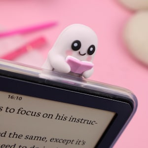 Op de afbeelding: Een wit spookvormig boekenlegger met een roze boek, geplaatst op een e-reader. De e-reader toont tekst en de tijd 16:10. De achtergrond is zachtroze.