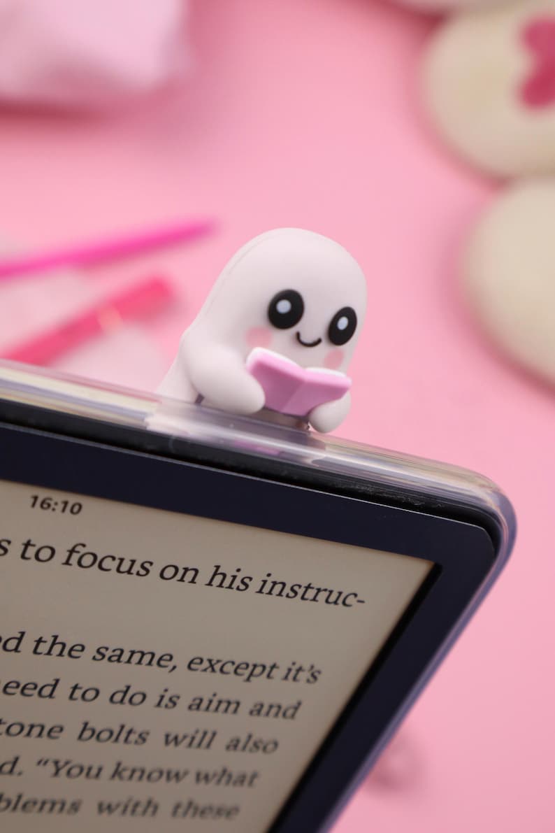 May include: A white ghost-shaped bookmark with a pink book, placed on top of an e-reader. The e-reader displays text, and the background is a soft pink. The bookmark has black eyes and pink cheeks.