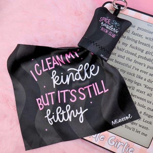 Op de afbeelding: Zwarte Kindle-hoes met de tekst "I clean my Kindle but it's still filthy" in roze en wit. Een kleinere zwarte pouch met "Spice Readers Book Club" is bevestigd. De achtergrond is roze.