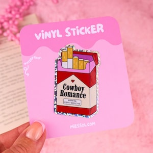 May include: A holographic vinyl sticker shaped like a cigarette pack with a red and white design. The pack features the text Cowboy Romance, a small red heart, and a warning label that reads WARNING: HIGHLY ADDICTIVE. The sticker is on a pink card.