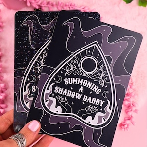 Könnte beinhalten: Zwei schwarze Ouija-Karten mit weißen und grauen Grafiken. Eine Karte zeigt den Text "SUMMONING A SHADOW DADDY" in einem Planchette-Design. Die andere Karte hat ein Sternennacht-Design. Die Karten werden vor einem rosa Hintergrund gehalten.
