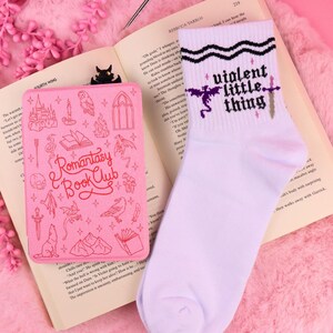 Könnte beinhalten: Ein pinkfarbener Romantasy Book Club Lesezeichen und ein Paar weiße Socken mit dem Aufdruck "violent little thing". Das Lesezeichen hat verschiedene Illustrationen, und die Socken zeigen ein Drachen- und Schwertdesign. Die Artikel werden auf einem offenen Buch präsentiert.