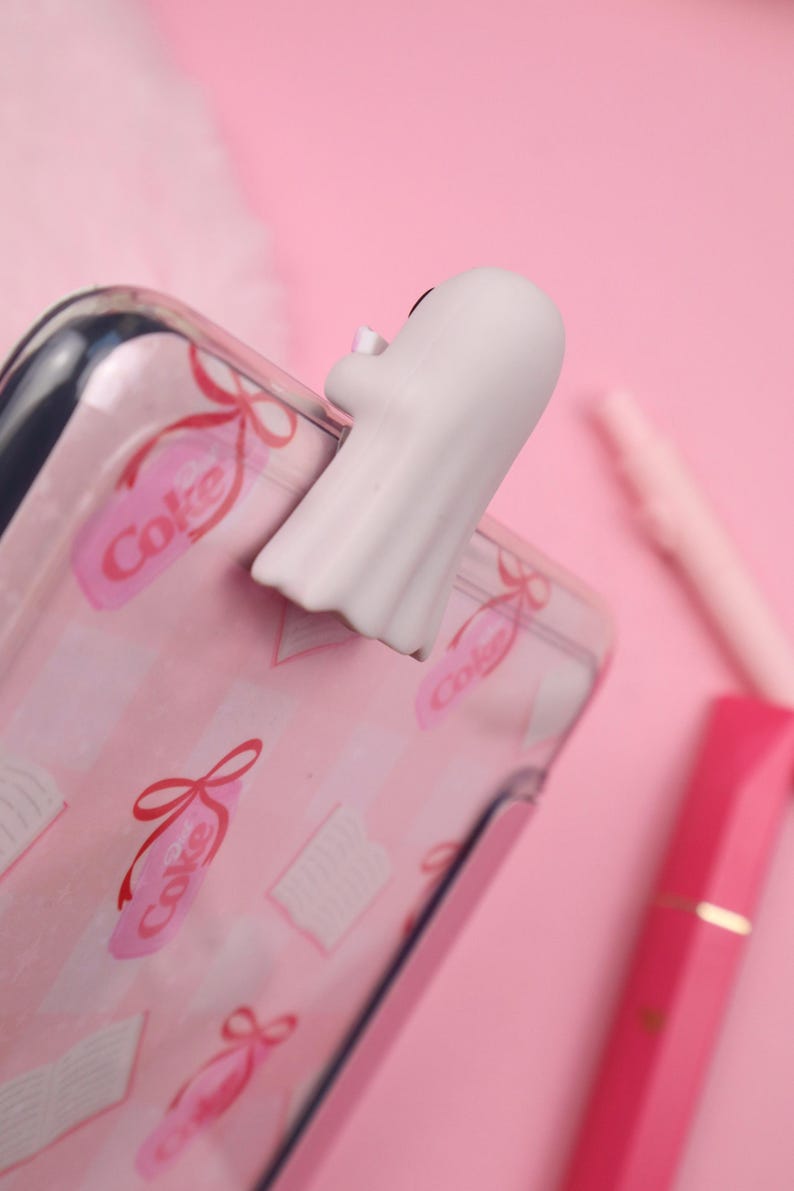 May include: A white ghost-shaped phone grip is attached to a pink phone case. The case has a pattern of pink ribbons, books, and the word "Coke". The background is a solid pink.