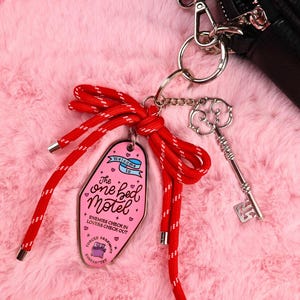 Puede incluir: Un llavero rosa estilo motel con el texto "The one bed Motel" y un cord&oacute;n rojo y blanco. Un dije de llave plateada y una cartera negra tambi&eacute;n son visibles. El llavero tiene el texto "Enemies check in, lovers check out."