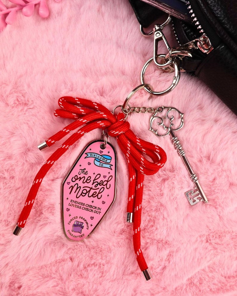 Puede incluir: Un llavero rosa estilo motel con el texto "The one bed Motel" y un cord&oacute;n rojo y blanco. Un dije de llave plateada y una cartera negra tambi&eacute;n son visibles. El llavero tiene el texto "Enemies check in, lovers check out."