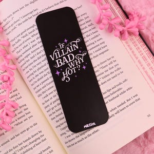 Könnte beinhalten: Schwarzer Lesezeichen mit dem Text "IF VILLAIN BAD WHY HOT?" in Weiß und Lila. Das Lesezeichen liegt auf einem offenen Buch mit rosa Blumen im Hintergrund. Das Lesezeichen hat abgerundete Ecken.