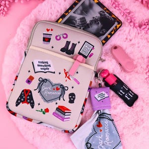 May include: A beige tablet case with a pocket, featuring illustrations of boots, a coffee cup, and the text "Dark Romance." A small pink device and a keychain with a lip gloss are also visible. The background is a pink, fluffy surface.