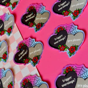 May include: Two heart-shaped stickers with a spiderweb design. One heart is black with the text "trigger warnings?" and the other heart is white with the text "yes please".