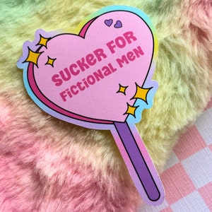 May include: A pink heart-shaped lollipop sticker with a rainbow border. The sticker says "SUCKER FOR FICTIONAL MEN" in white text. There are small yellow stars and hearts around the text.