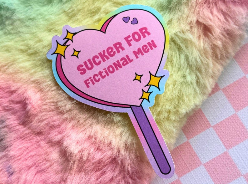 May include: A pink heart-shaped lollipop sticker with a rainbow border. The sticker says "SUCKER FOR FICTIONAL MEN" in white text. There are small yellow stars and hearts around the text.