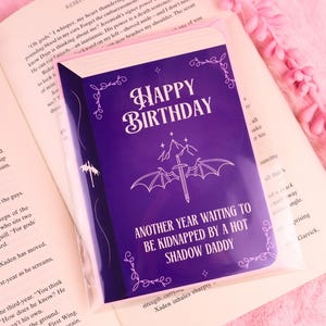Könnte beinhalten: Eine lila Geburtstagskarte mit dem Text "HAPPY BIRTHDAY" und "ANOTHER YEAR WAITING TO BE KIDNAPPED BY A HOT SHADOW DADDY". Die Karte befindet sich in einer transparenten Plastikhülle und liegt auf einem offenen Buch.