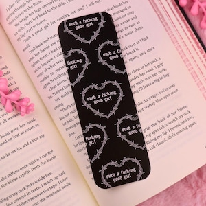 Op de afbeelding: Zwarte boekenlegger met witte hartontwerpen van prikkeldraad en de zinnen "such a fucking good girl" en "fucking girl". De boekenlegger ligt op een open boek met roze accenten.