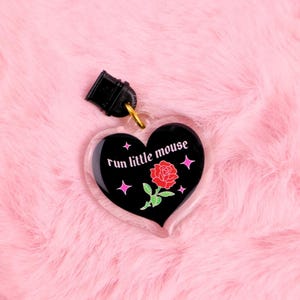 K&ouml;nnte beinhalten: Ein herzf&ouml;rmiger Schl&uuml;sselanh&auml;nger mit dem Text "run little mouse" in wei&szlig;er Schrift. Das schwarze Herz zeigt eine rote Rosenillustration und rosa Sternakzente. Der Schl&uuml;sselanh&auml;nger hat einen schwarzen Verschluss und einen goldfarbenen Ring.