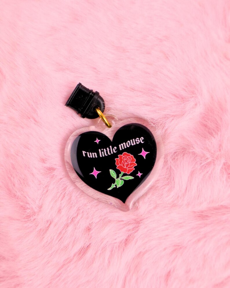 K&ouml;nnte beinhalten: Ein herzf&ouml;rmiger Schl&uuml;sselanh&auml;nger mit dem Text "run little mouse" in wei&szlig;er Schrift. Das schwarze Herz zeigt eine rote Rosenillustration und rosa Sternakzente. Der Schl&uuml;sselanh&auml;nger hat einen schwarzen Verschluss und einen goldfarbenen Ring.