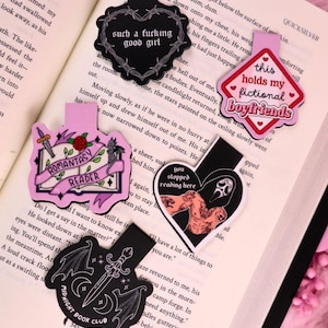 May include: A selection of decorative bookmarks with diverse designs and text, such as "such a fucking good girl" and "this holds my fictional boyfriends." The bookmarks are in black, pink, and white, featuring illustrations and text.