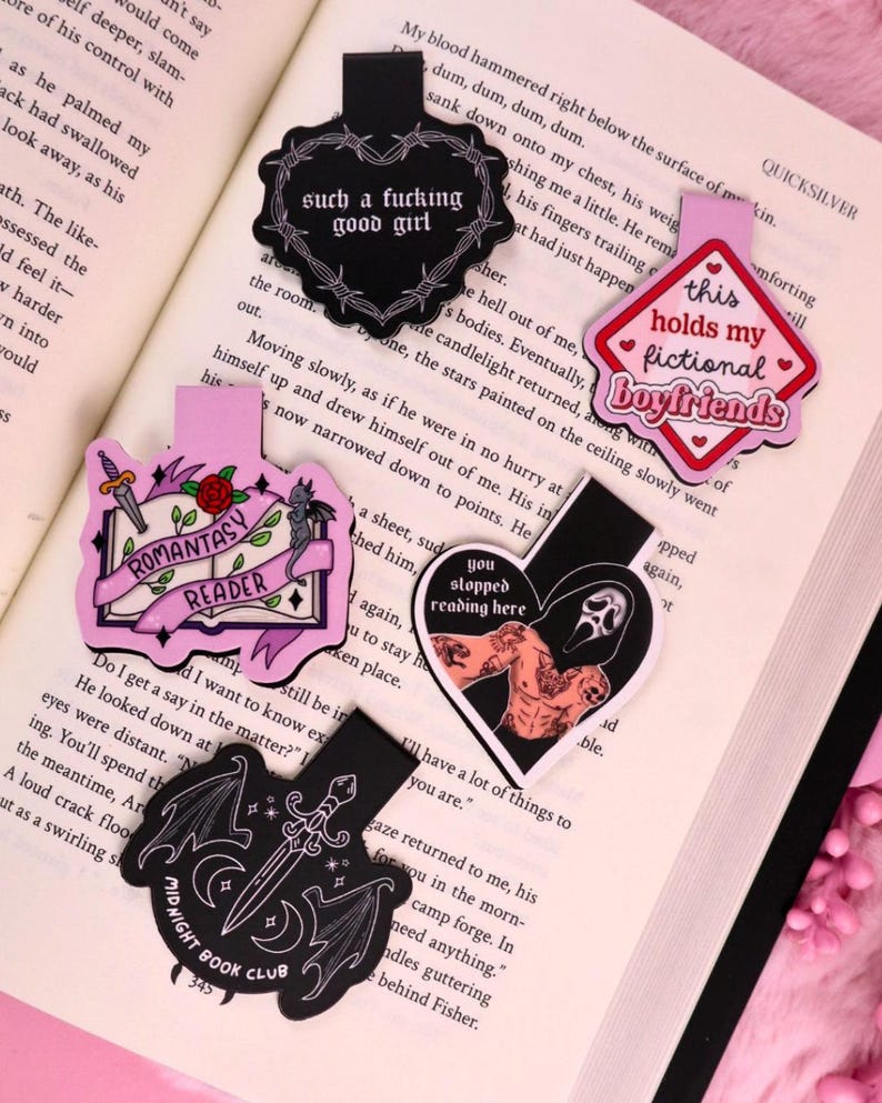 May include: A selection of decorative bookmarks with diverse designs and text, such as "such a fucking good girl" and "this holds my fictional boyfriends." The bookmarks are in black, pink, and white, featuring illustrations and text.