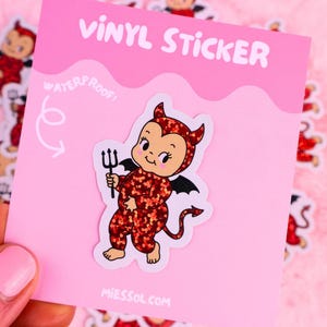 Op de afbeelding: Een roze vinyl sticker met een cartoon duivelsbaby met rode glitterdetails, die een drietand vasthoudt. De sticker zit op een roze kaart met de woorden "VINYL STICKER" en "WATERPROOF!" erop gedrukt. Het websiteadres MIESSOL.COM is ook zichtbaar.