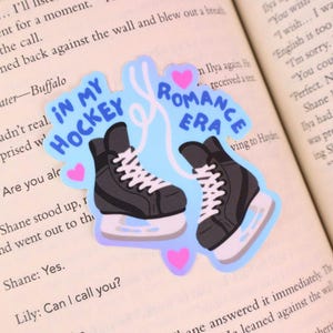 May include: A sticker featuring two black ice skates with white laces and blades, set against a light blue background. The text "IN MY HOCKEY ROMANCE ERA" is written in blue. Pink hearts accent the design.