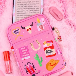 Könnte beinhalten: Eine rosa Tablethülle mit verschiedenen Western-Motiven, darunter ein Kuhschädel, ein Hufeisen, ein Kaktus, ein Cowboyhut, ein rosa Stiefel und eine Zigarettenschachtel mit der Aufschrift Cowboy Romance sowie Texten wie Atta Girl und Boots Stay On.