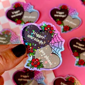 May include: Two holographic heart-shaped stickers with the text "trigger warnings?" and "yes please" in a gothic font. The stickers are on a pink and white checkered background.
