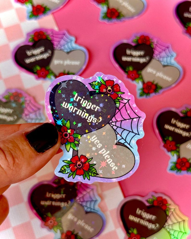 May include: Two holographic heart-shaped stickers with the text "trigger warnings?" and "yes please" in a gothic font. The stickers are on a pink and white checkered background.