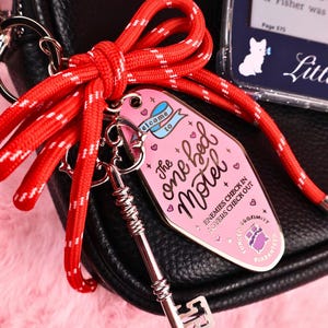 Puede incluir: Un llavero rosa con forma de llave de motel con el texto "The one bed Motel" y una llave plateada sujeta a una cuerda roja y blanca. La etiqueta tiene un dise&ntilde;o vintage con corazones y la frase "Enemies check in, lovers check out."