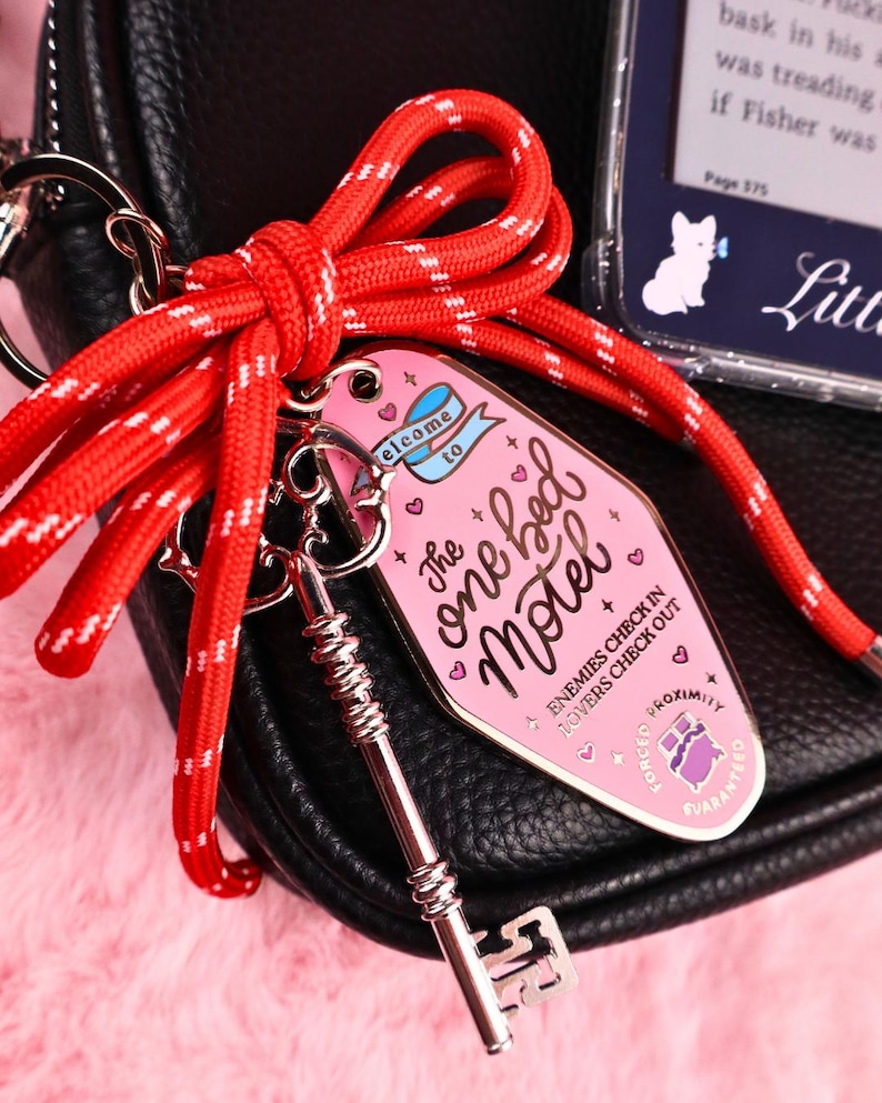 Puede incluir: Un llavero rosa con forma de llave de motel con el texto "The one bed Motel" y una llave plateada sujeta a una cuerda roja y blanca. La etiqueta tiene un dise&ntilde;o vintage con corazones y la frase "Enemies check in, lovers check out."