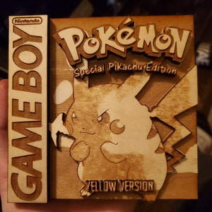 Könnte beinhalten: Ein Holzausschnitt einer Nintendo Game Boy-Spielkassette für das Spiel Pokemon Special Pikachu Edition Yellow Version. Der Ausschnitt ist braun und zeigt ein gelbes Pikachu mit schwarzem Umriss.
