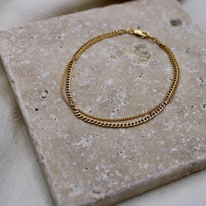 May include: A gold chain bracelet with two different chain styles. The bracelet is on a light brown textured surface.