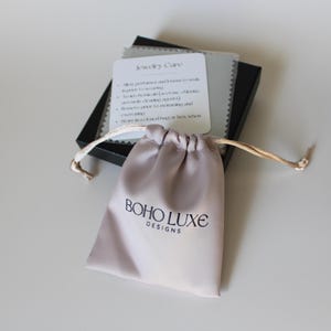 May include: A light gray satin jewelry pouch with a drawstring closure. The pouch has the text "BOHO LUXE DESIGNS" printed in black.
