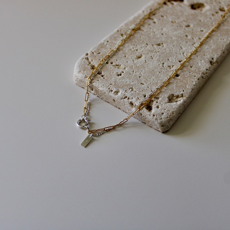 May include: A silver and gold chain necklace with a small rectangular pendant. The necklace is on a light brown textured surface.
