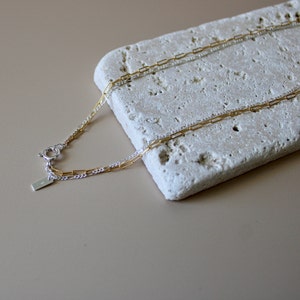 May include: A gold and silver chain necklace with a clasp closure. The necklace is on a white textured surface.