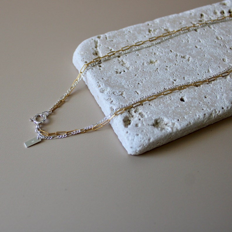 May include: A gold and silver chain necklace with a clasp closure. The necklace is on a white textured surface.
