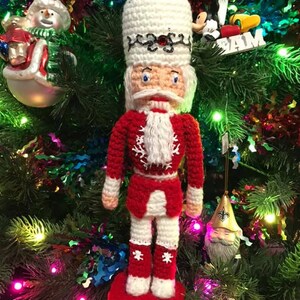 May include: A crocheted nutcracker ornament in red, white, and black with a white beard and a red hat. The ornament is hanging on a Christmas tree with other ornaments.