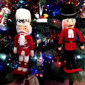 May include: Two crocheted nutcracker ornaments hanging on a Christmas tree. One nutcracker is wearing a red and white outfit with a white hat, while the other is wearing a red and black outfit with a black hat. Both nutcrackers have white beards and are smiling.