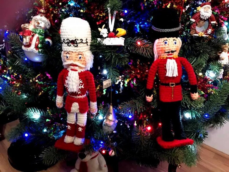 May include: Two crocheted nutcracker ornaments hanging on a Christmas tree. One nutcracker is wearing a red and white outfit with a white hat, while the other is wearing a red and black outfit with a black hat. Both nutcrackers have white beards and are smiling.