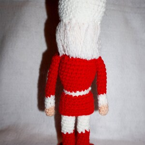 May include: A crocheted nutcracker doll with a red and white outfit. The doll is standing on a red base.
