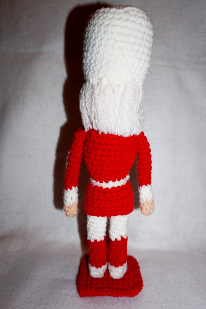 May include: A crocheted nutcracker doll with a red and white outfit. The doll is standing on a red base.