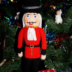 May include: A crocheted nutcracker doll wearing a black hat, red coat, and black pants. The doll is standing on a red and white gift box with a snowman pattern.