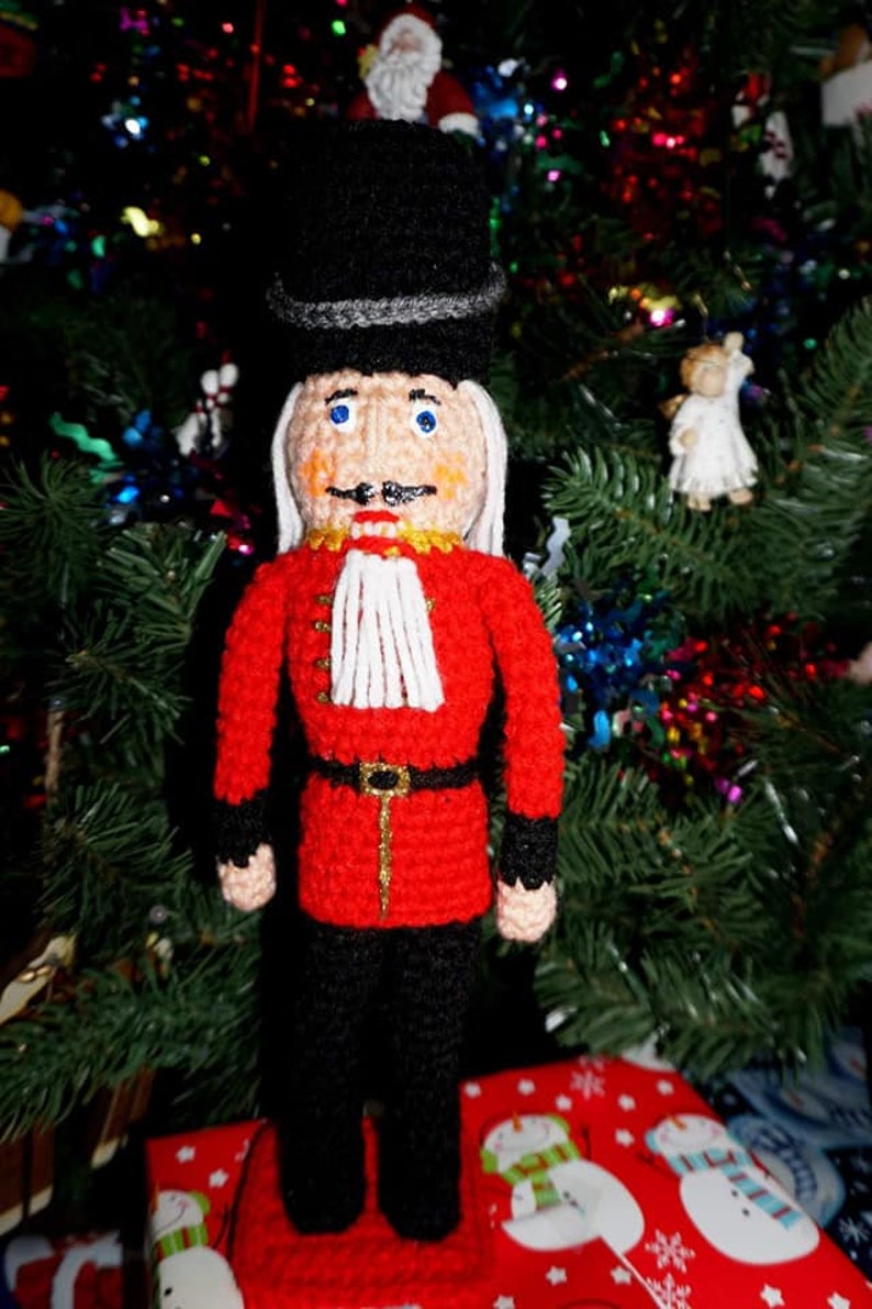 May include: A crocheted nutcracker doll wearing a black hat, red coat, and black pants. The doll is standing on a red and white gift box with a snowman pattern.