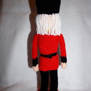 May include: A crocheted toy soldier wearing a red uniform, black pants, and a black hat with a white beard. The soldier is standing on a red base.