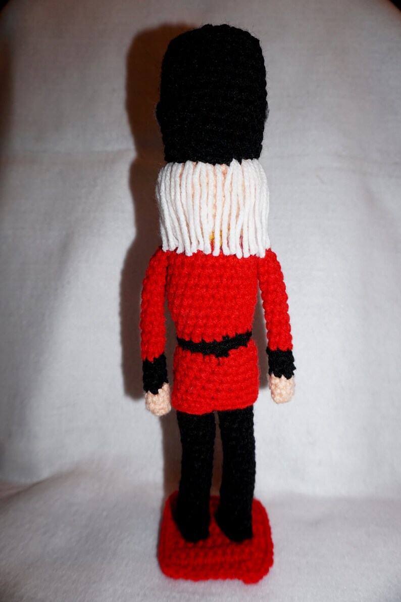 May include: A crocheted toy soldier wearing a red uniform, black pants, and a black hat with a white beard. The soldier is standing on a red base.
